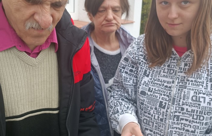 Starszy mężczyzna w czapce i czarno-czerwonej kurtce trzyma wiklinowy koszyk wypełniony foliowymi torebkami. Za nim stoją dwie osoby, jedna z nich ma szarą kurtkę z wzorem róży