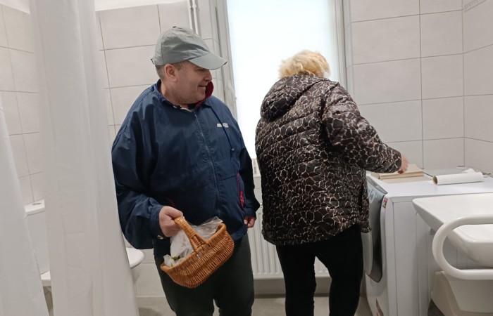 Mężczyzna w granatowej kurtce trzyma koszyk, stojąc w jasnym pomieszczeniu przypominającym łazienkę. Kobieta w czarnej kurtce stoi przy pralce, przeszukując blat. W tle białe płytki i umywalka