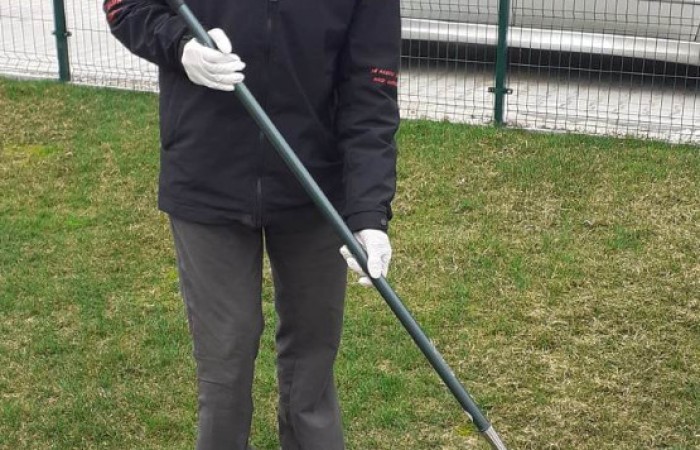 Starszy mężczyzna w czarno-czerwonej kurtce, czarnej czapce i białych rękawiczkach grabi trawnik za pomocą szerokich metalowych grabi. W tle widoczne są ogrodzenie z siatki, biały samochód i budynek z pochylonym dachem.