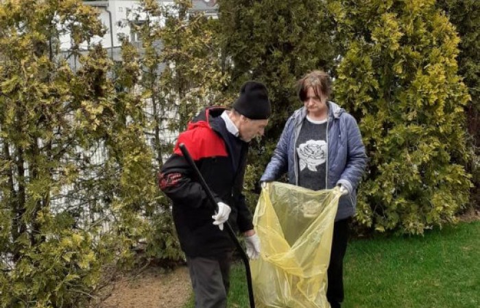 Dwie osoby wykonują prace ogrodowe na trawniku. Mężczyzna w czapce i czarno-czerwonej kurtce trzyma grabie i zbiera liście, a kobieta w niebieskiej kurtce trzyma otwarty worek na odpady ogrodowe w kolorze żółtym. W tle widoczne są tuje oraz słup oświetleniowy.
